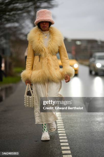 Guest wears a pale pink fluffy bob hat, a white latte turtleneck pullover, a yellow shiny leather with embroidered fluffy borders, a beige long...