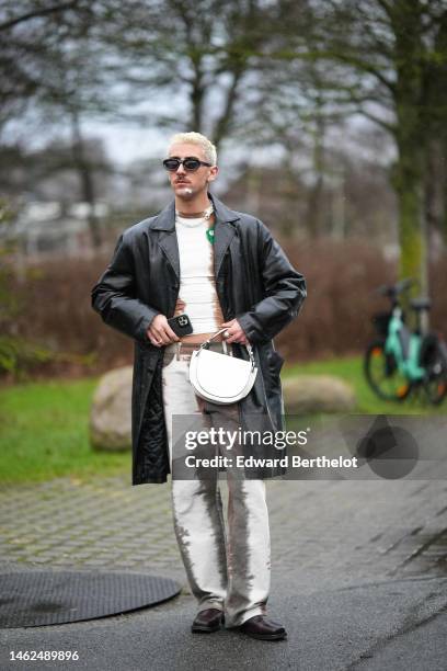 Guest wears black sunglasses, silver earrings, a silver large chain necklace, a white and brown tie and dye print denim large pants, a black shiny...