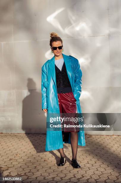 Mija Knezevic wearing a blue varnished shiny leather coat, burgundy shiny leather shorts, black tights, light blue top, and navy cardigan outside...