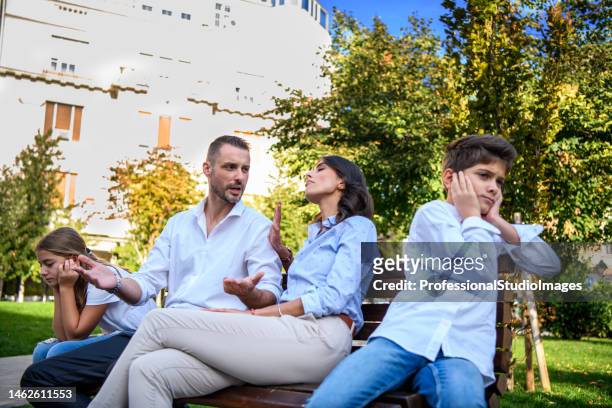 handsome man e sua moglie hanno una discussione seria mentre sono seduti sulla panchina nel parco cittadino con i bambini. - diverbio foto e immagini stock