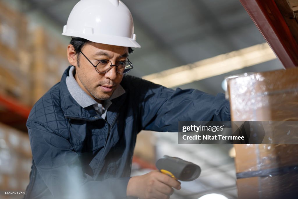 Warehouse Technology and Automation in Supply Chain Management. Low-angle view of a male Japanese warehouse leader used a barcode reader to scan a product code and confirm product picking in a factory distribution warehouse.