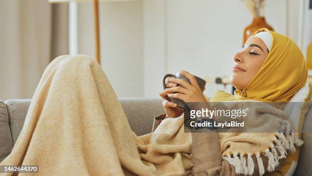 entspannen sie sich, sofa und frau im hijab trinken kaffee mit lächeln und decke gemütlich im wohnzimmer liegend. entspannende teezeit, islamische person, die auf der couch chillt und in ruhe und frieden in der home lounge denkt - arabische frau kopftuch stock-fotos und bilder