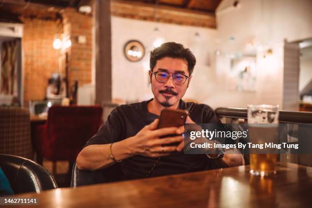 young handsome asian man using smartphone while enjoying beer in pub at night - modern manhood - fotografias e filmes do acervo