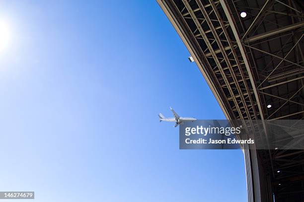 a jet airliner flying past an open hangar door at an airport. - sydney airport foto e immagini stock