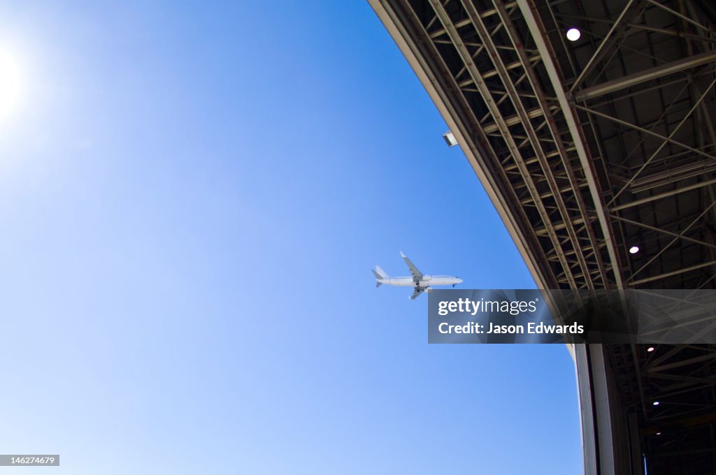 A jet airliner flying past an open hangar door at an airport.