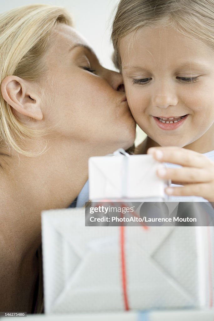 Mother kissing young daughter's cheek as girl looks at wrapped gifts