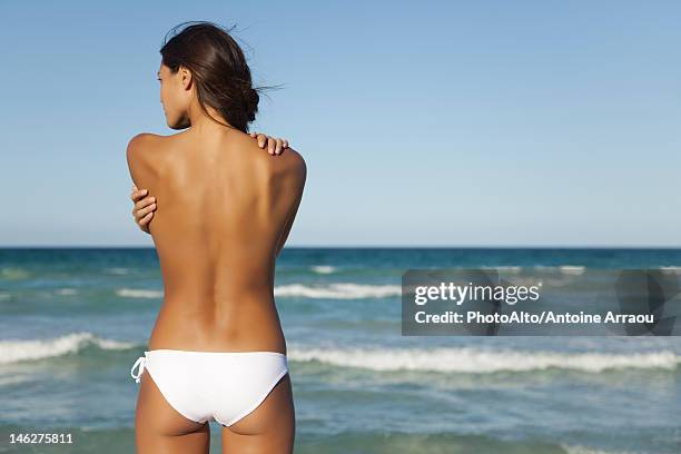 semi-naked woman in bikini bottom hugging self by ocean, rear view - beach bum stock pictures, royalty-free photos & images