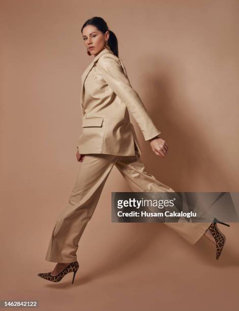 masculine-looking beautiful woman in a leather suit is jumping in front of a brown background. - modieus stockfoto's en -beelden