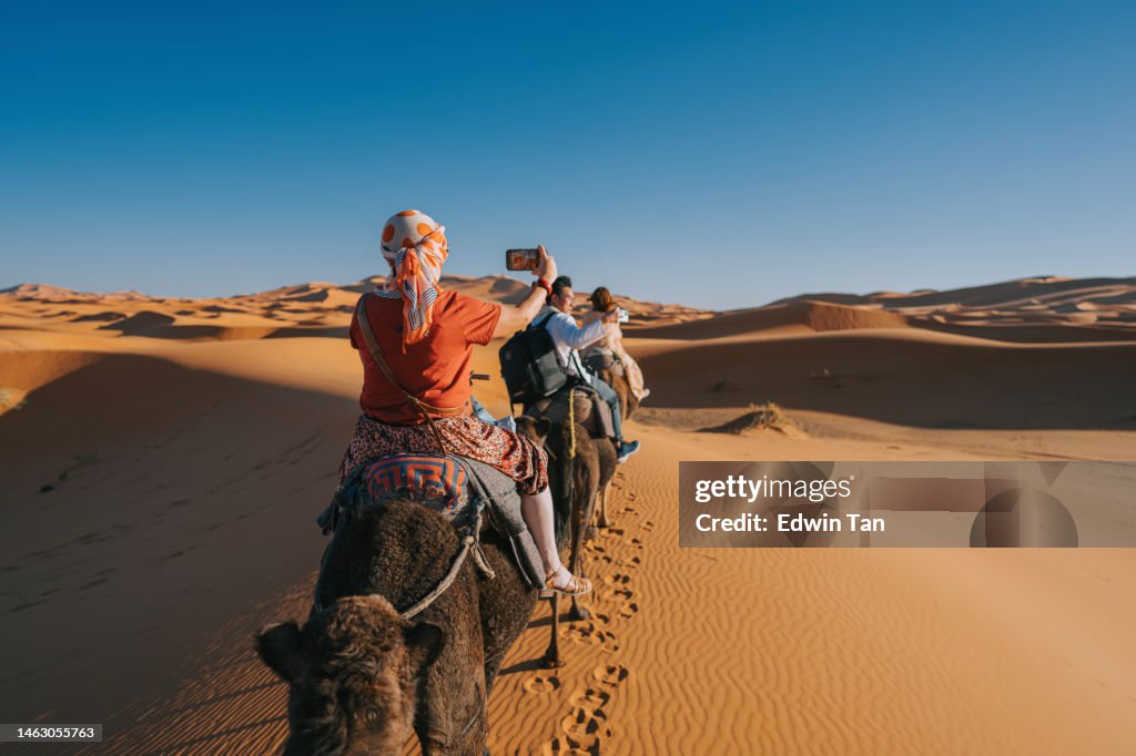 Turistas chineses asiáticos que andam de trem de camelo dromedário cruzando o deserto do Saara Marrocos liderado por pastor guia turístico durante o pôr do sol