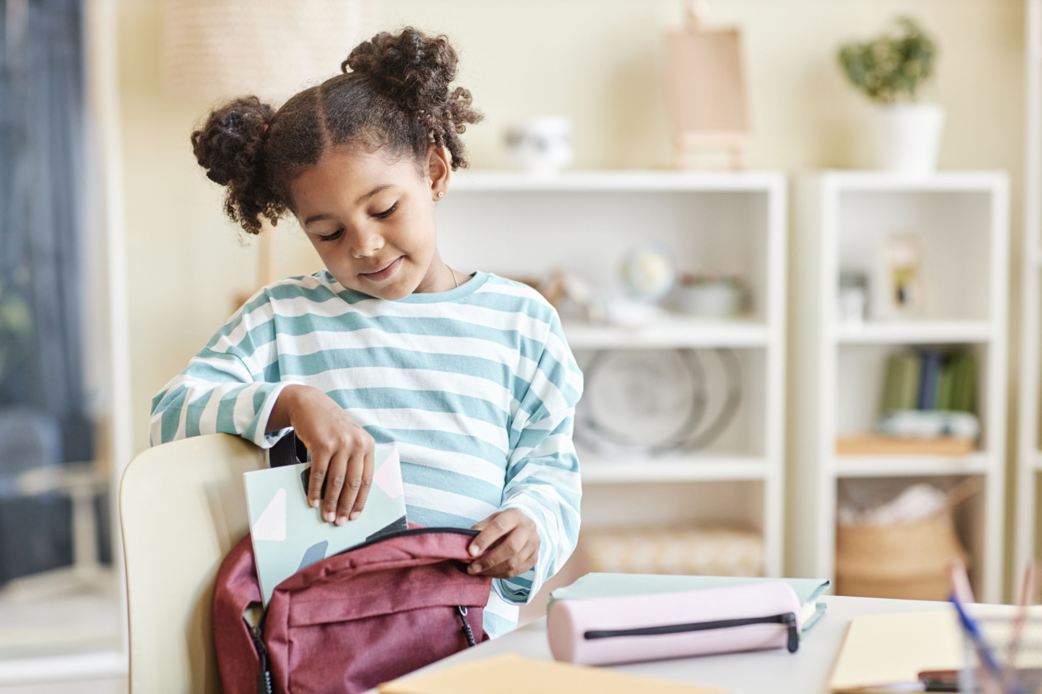 Cute black girl packing backpack for school in pastel home interior Cute black girl packing backpack for school in pastel home interior