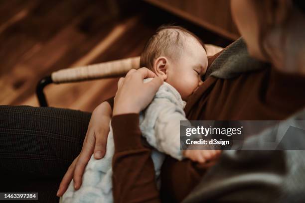 over the shoulder view of a loving young asian mother sitting on the armchair in the living room, breastfeeding her baby at home. family moments, love, care and togetherness concept - birthing chair stock pictures, royalty-free photos & images