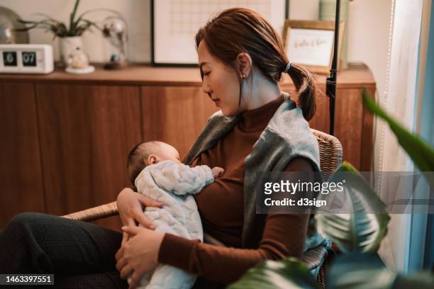 a loving young asian mother sitting on the armchair in the living room, breastfeeding her baby at home. family moments, love, care and togetherness concept - birthing chair stock pictures, royalty-free photos & images