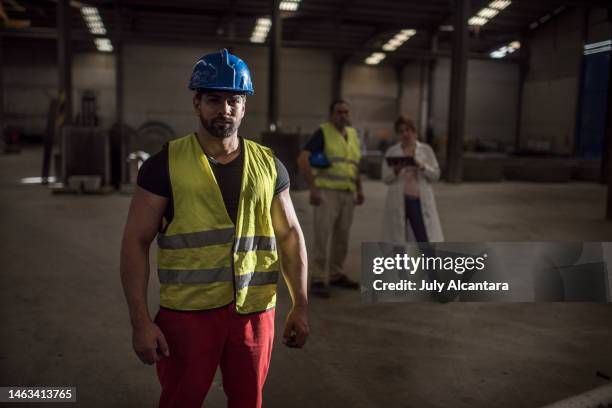 trabajador de la construcción posa con una iluminación dramática mirando a la cámara con sus compañeros de trabajo en el fondo (hombre y mujer de laboratorio embarazada) - pregnant july 2017 fotografías e imágenes de stock