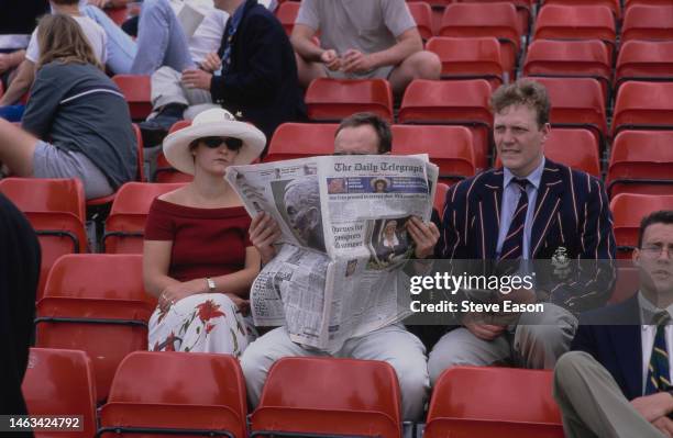 Woman and two men sitting watching the Henley Regatta, one man is reading a copy of The Daily Telegraph newspaper, the other is dressed in a striped...