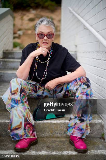 portrait, cool and funky mature woman with stylish and trendy fashion sitting on stairs outdoors. old, elderly and urban outfit by pensioner female with modern and colorful clothes on steps - anti ageing stockfoto's en -beelden
