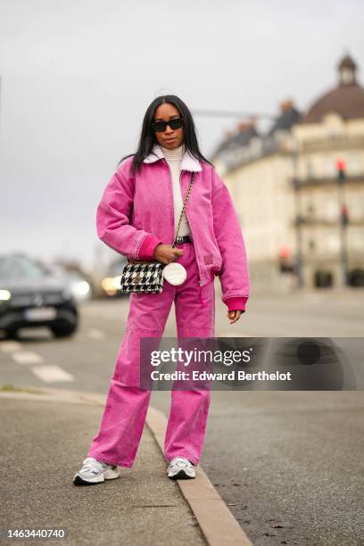 Guest wears black sunglasses, a white turtleneck pullover, a pink denim zipper bomber coat, matching pink denim wide legs pants, a black and white...