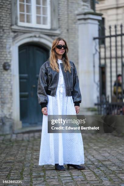 Guest wears black sunglasses, a white cut-out polka dots pattern buttoned / belted long dress, a black faded leather bomber coat, black shiny leather...