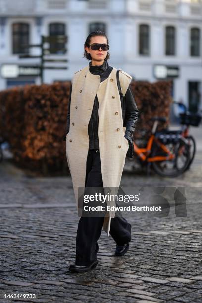 Guest wears black futurist sunglasses, a black shiny leather turtleneck jacket, a beige checkered print pattern sleeveless / buttoned long coat,...