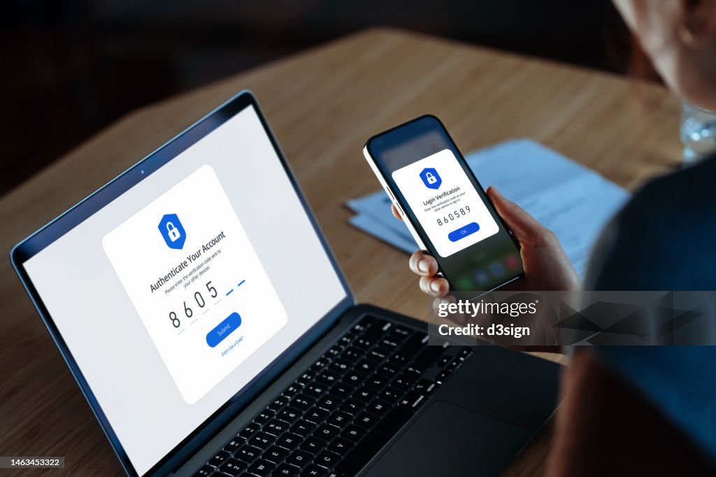 Over the shoulder view of young woman using mobile device with Two-Factor Authentication (2FA) security while logging in securely to her laptop. Privacy protection, internet and mobile security