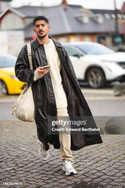 Guest wears a white wool pullover, beige denim pants, a black shiny leather oversized long coat, a white latte shiny leather handbag, white leather...