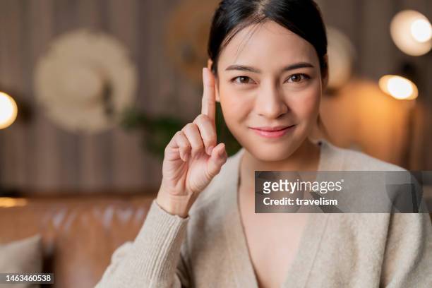 porträt der asiatischen frau attraktive frau blick auf die kamera hand finger zeigen nachdenkliche emotion gesichtsausdruck zu hause - one finger selfie stock-fotos und bilder