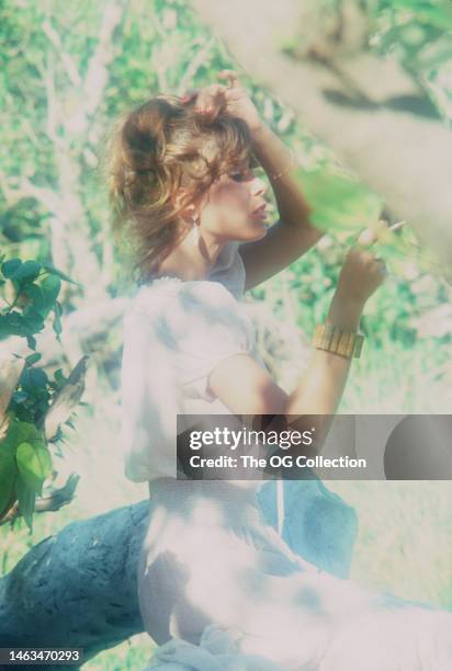 American model and actress Corinne Alphen holds her hair in one hand and a cigarette in the other, as she poses among trees outdoors, May 1981. The...