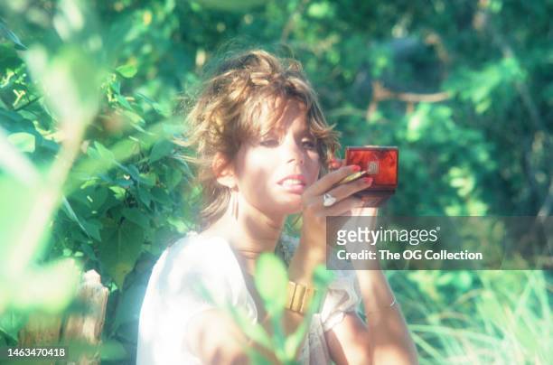 American model and actress Corinne Alphen looks at her make-up compact as she poses among trees outdoors, May 1981. The photo was taken as part of...