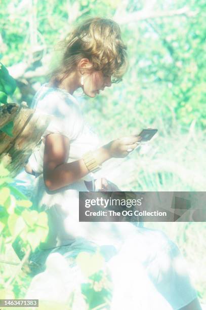 American model and actress Corinne Alphen looks at her make-up compact as she poses among trees outdoors, May 1981. The photo was taken as part of...