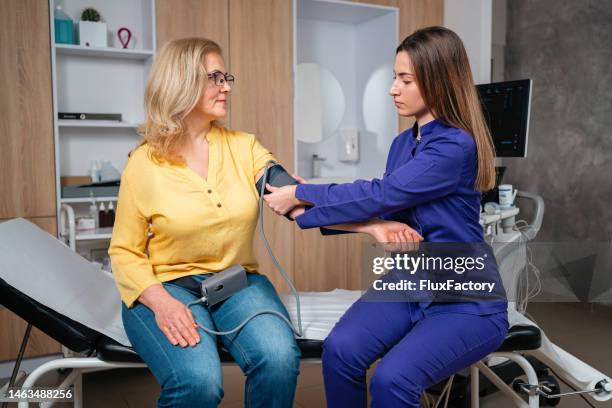 une patiente âgée assise sur une table d’examen pendant qu’une infirmière de race blanche mesure la tension artérielle - table dexamen médical photos et images de collection