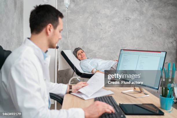 male doctor working on a computer at the doctor's office, while a senior female patient getting an infusion - vitamin iv stock pictures, royalty-free photos & images