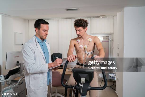 male patient with electrodes on his chest, doing stress test on an exercise bicycle, while doctor observing him - stress test stock pictures, royalty-free photos & images