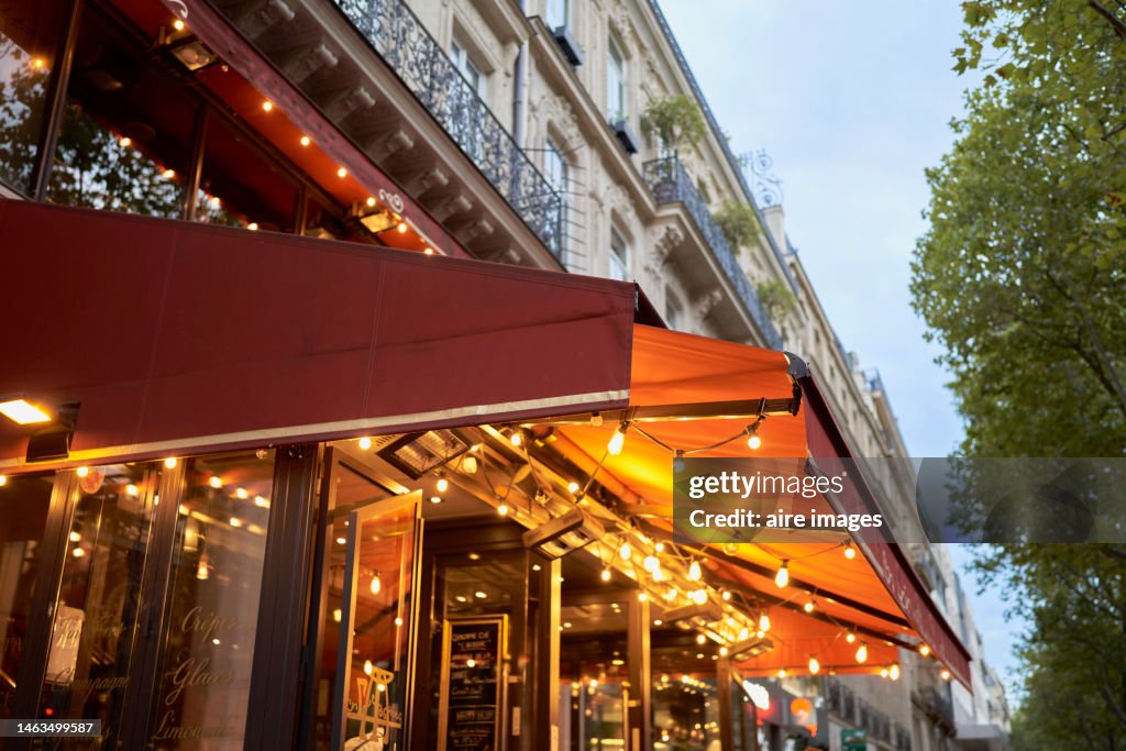 Facade of a cafeteria with a red awning decorated with lights, on the upper part there are apartments with balconies and in one there is a flower pot.