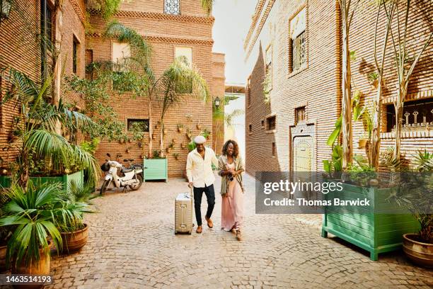 wide shot of couple arriving in hotel courtyard with rolling luggage - suitcase couple stockfoto's en -beelden