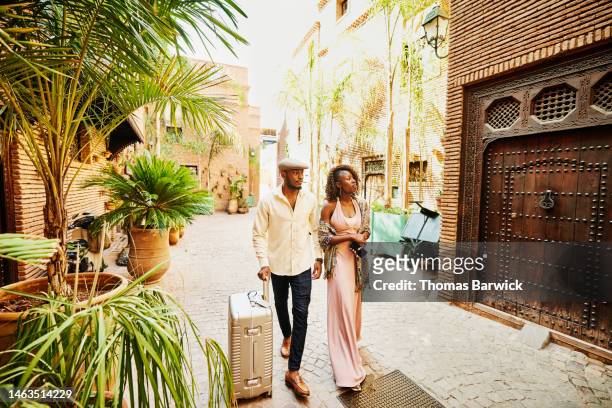 Wide shot of couple arriving in hotel courtyard with rolling luggage