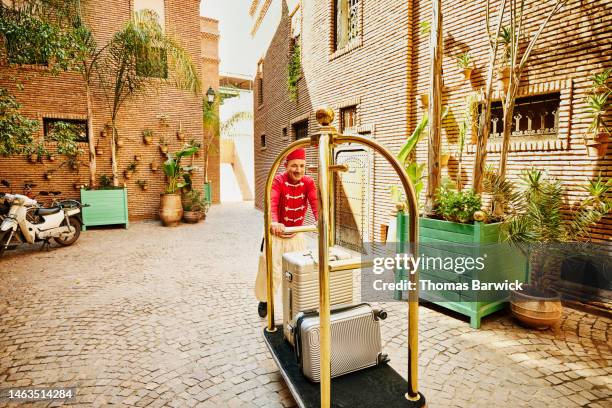 wide shot of smiling bellman moving guests luggage in hotel courtyard - bell boy stock pictures, royalty-free photos & images