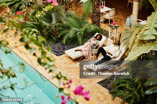 Wide shot of couple relaxing in lounge chairs by pool at luxury hotel