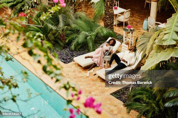 wide shot of couple relaxing in lounge chairs by pool at luxury hotel - travel and not business stock pictures, royalty-free photos & images