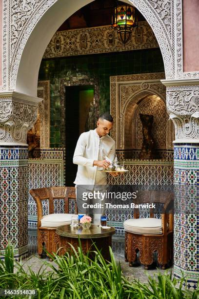 Wide shot of waiter preparing tea service in courtyard of luxury hotel