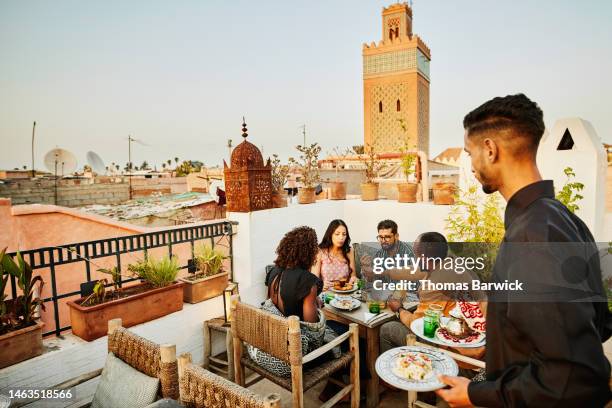 wide shot of waiter bringing dishes to friends having rooftop dinner - rooftop dining foto e immagini stock