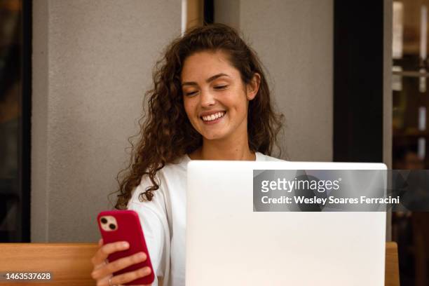 young adult woman with long curly hair sitting on a wooden table with her laptop and checking a cell phone with red cover - phone cover stockfoto's en -beelden