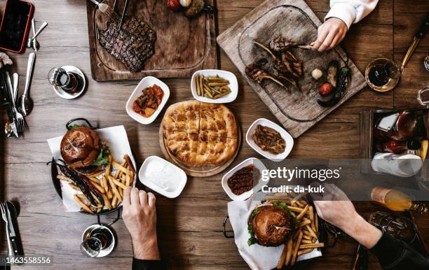 draufsicht auf rindersteaks, burger und snacks mit gegrilltem gemüse auf dem tisch. menschen, die verschiedene lebensmittel zu sich nehmen - steakhouse stock-fotos und bilder