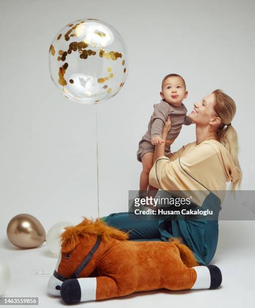 baby boy in mother's lap looking at her flying balloon in front of white background. - helium stock pictures, royalty-free photos & images
