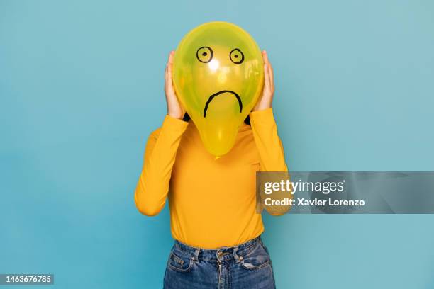 young woman hidden behing a balloon with a sad face drawn on it over blue background. negative emotion concept - faute photos et images de collection