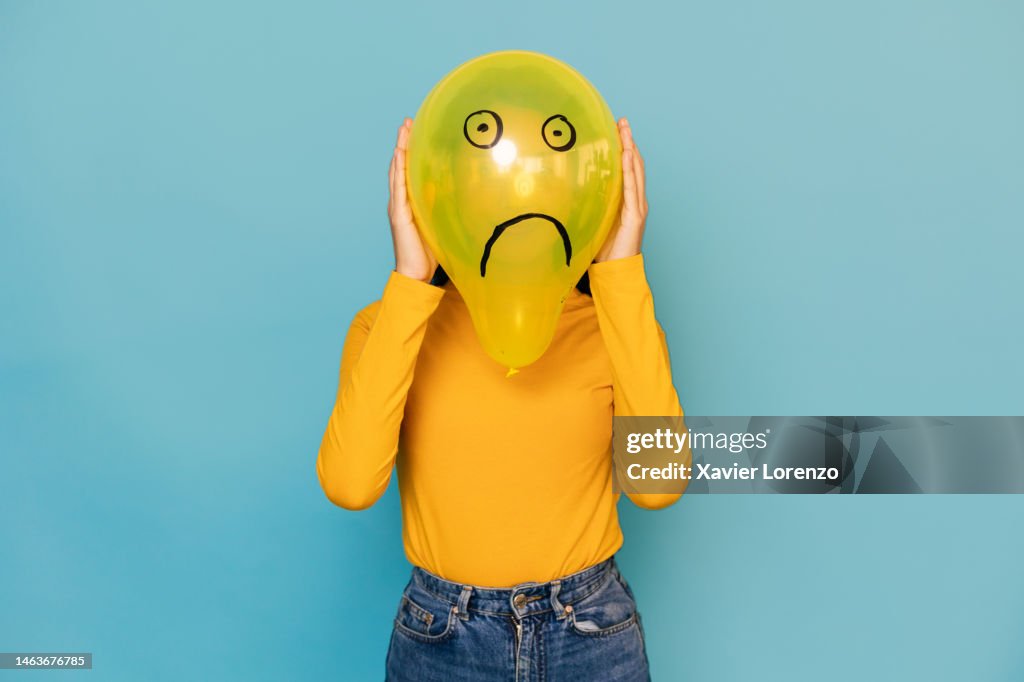 Young woman hidden behing a balloon with a sad face drawn on it over blue background. Negative emotion concept