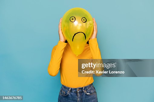Young woman hidden behing a balloon with a sad face drawn on it over blue background. Negative emotion concept Young woman hidden behing a balloon with a sad face drawn on it over blue background. Negative emotion concept