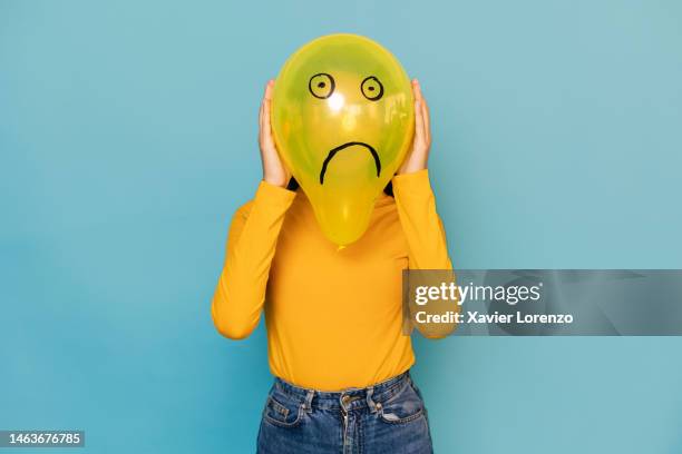 young woman hidden behing a balloon with a sad face drawn on it over blue background. negative emotion concept - stress emotivo imagens e fotografias de stock