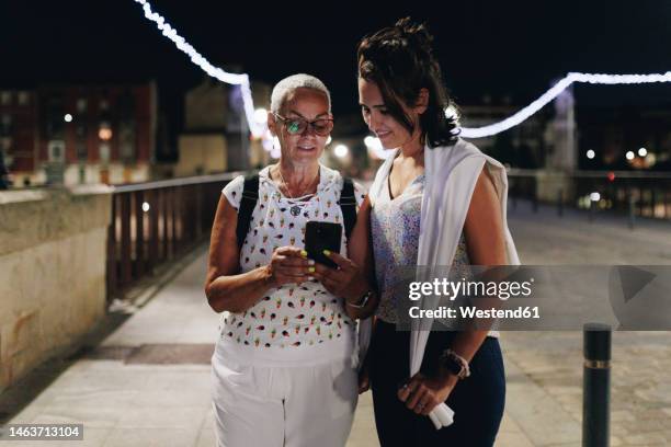senior woman sharing mobile phone with friend on footpath at night - zuid europese etniciteit stockfoto's en -beelden