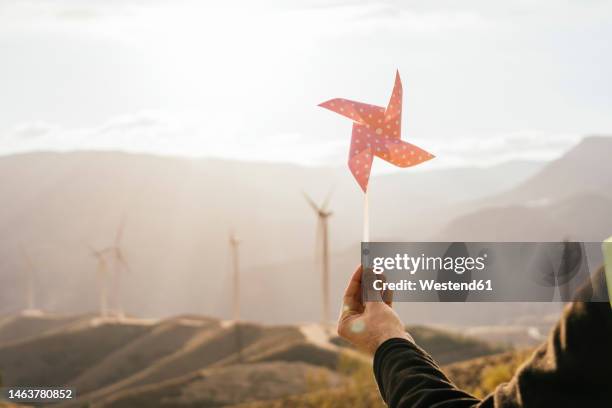 hand of technician holding pinwheel toy in front of wind turbines - paper windmill stock-fotos und bilder