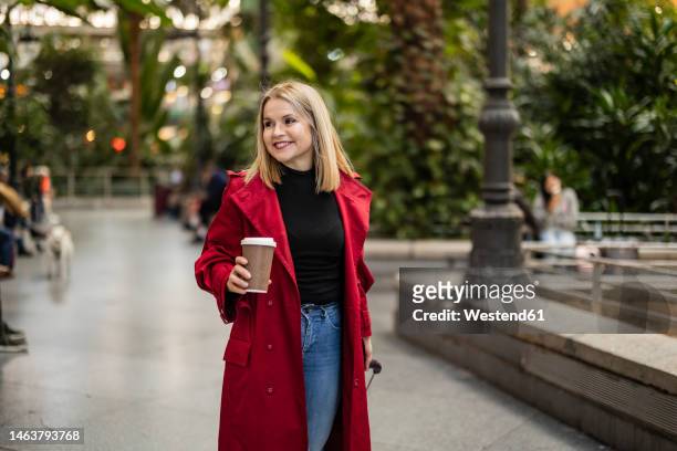 smiling mature woman holding disposable coffee cup on footpath - red coat stock pictures, royalty-free photos & images