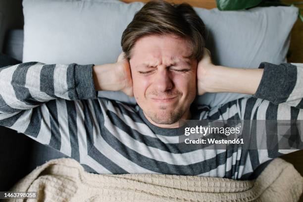 frustrated man covering ears lying on bed at home - hands covering ears stock pictures, royalty-free photos & images